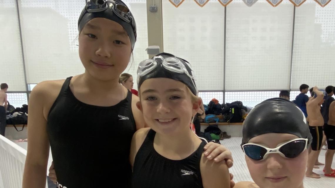 Three young swimmers, all in caps and one wearing goggles, pose together by the pool