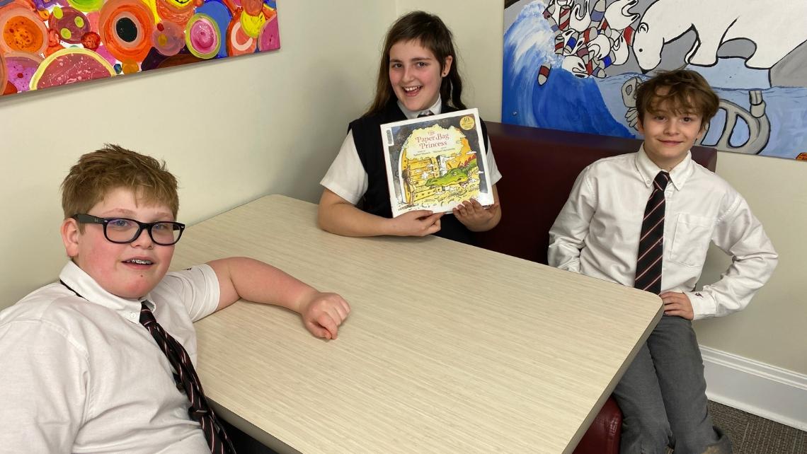 Three students seated at a table, one is holding the book The Paper Bag Princess by Robert Munsch